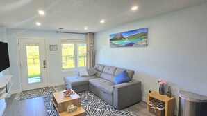 Ein modernes Wohnzimmer mit einem grauen Ecksofa, einem Flachbildfernseher, einem Gemälde von einem Bergsee und einem Holztisch.