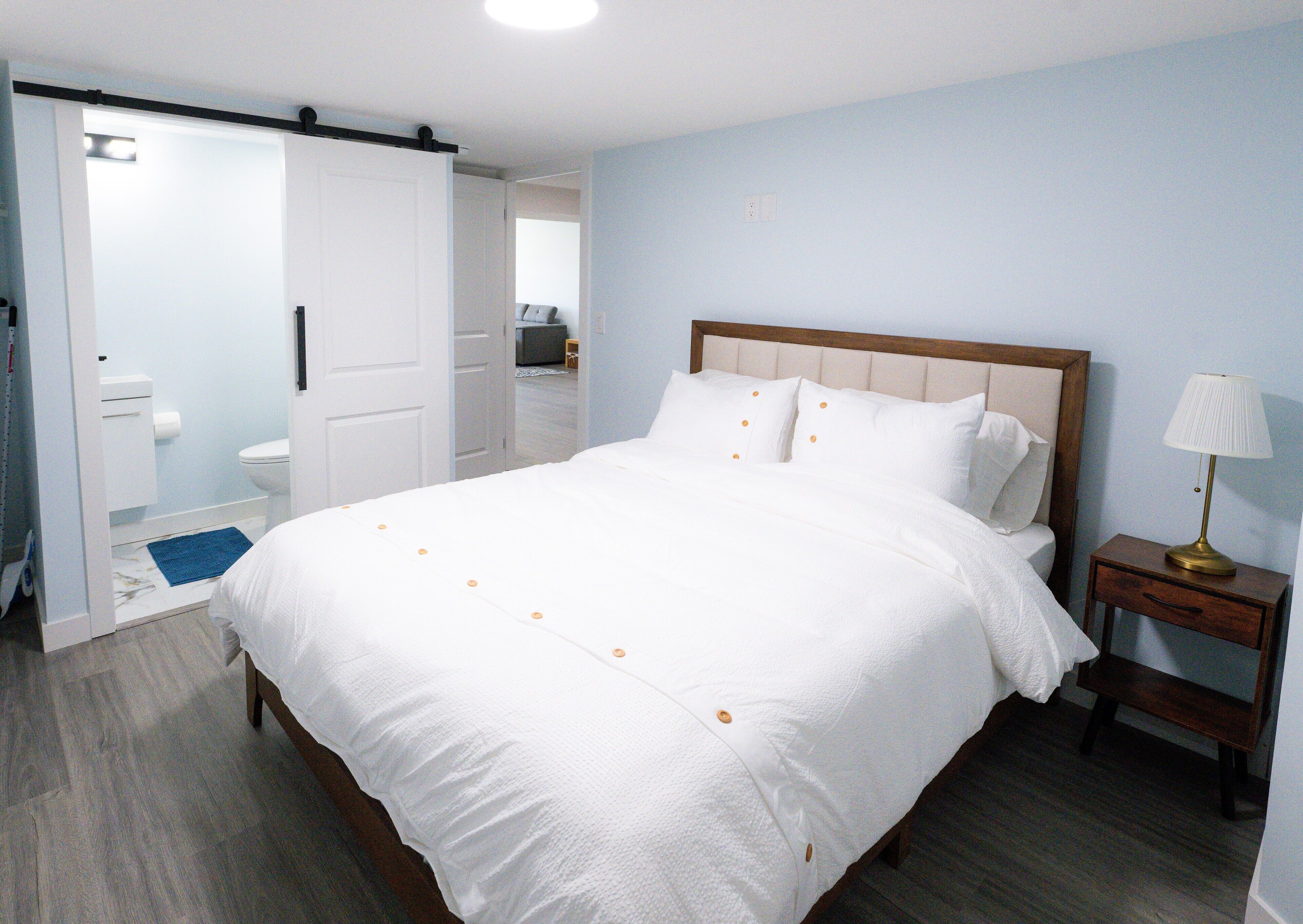 A neatly made bed with white bedding and orange buttons, a wooden nightstand with a lamp, and a bathroom visible through an open door.