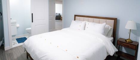 A neatly made bed with white bedding and orange buttons, a wooden nightstand with a lamp, and a bathroom visible through an open door.