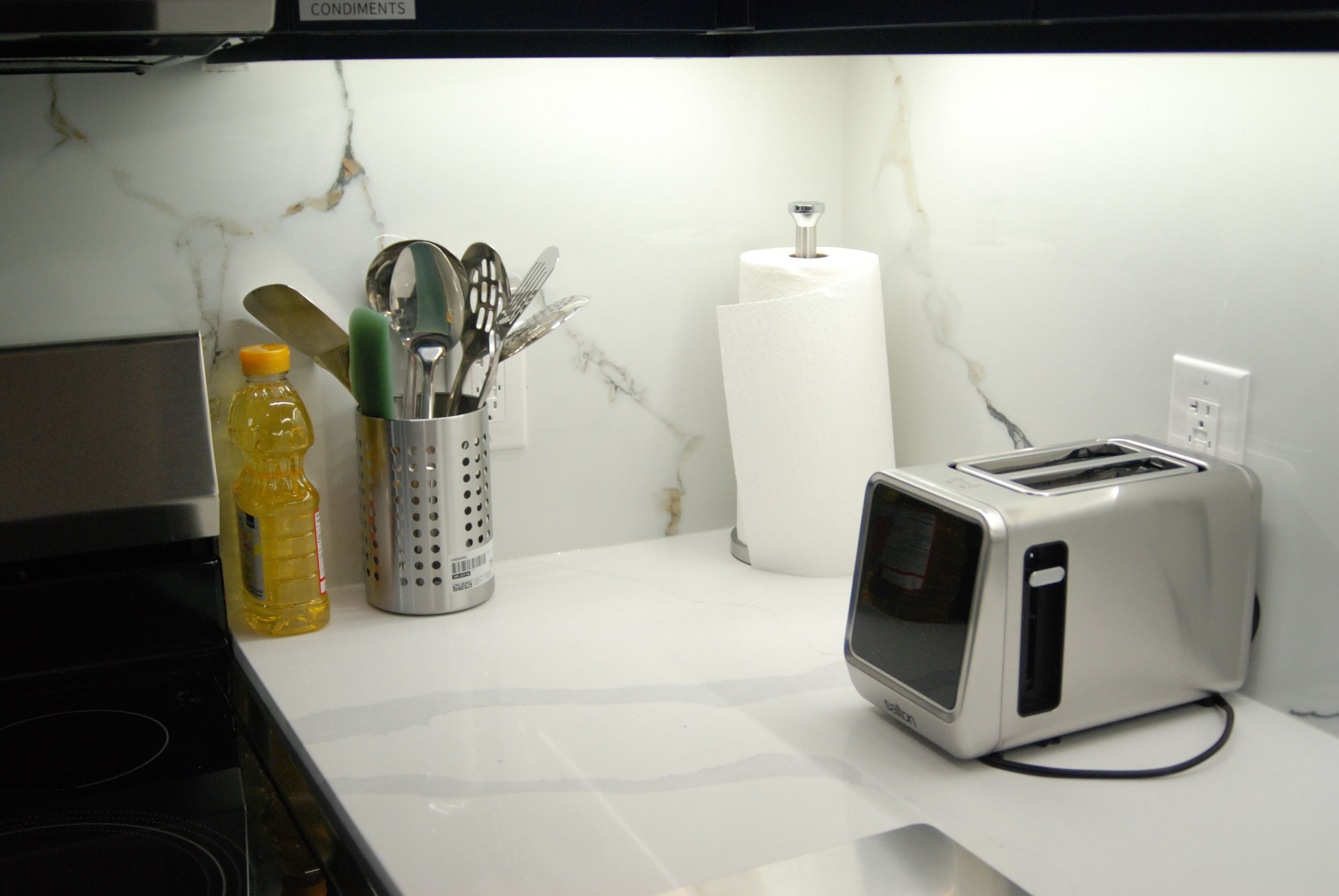 A kitchen counter with a toaster, a bottle of yellow liquid, a container with utensils, and a roll of paper towels.