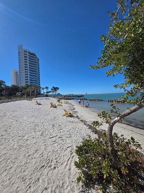 A beach with sand, a few yellow lounge chairs, and a tall building in the background. - Sarasota Stunner Apartment. Prime Location, Beautiful Design. (Sarasota)