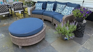 Un espace patio aménagé avec un canapé en osier, des coussins bleus et un pouf rond. On y trouve également des plantes en pot et une petite table avec des chaises.