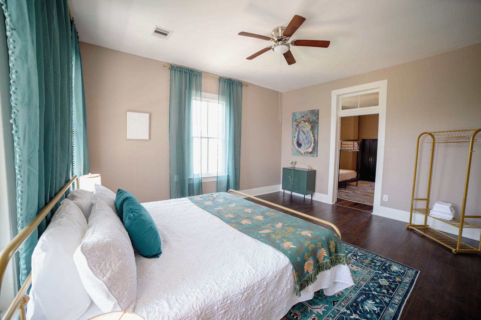 A bedroom with a bed, a ceiling fan, a window with curtains, a small table, a chair, and a walk-in closet.