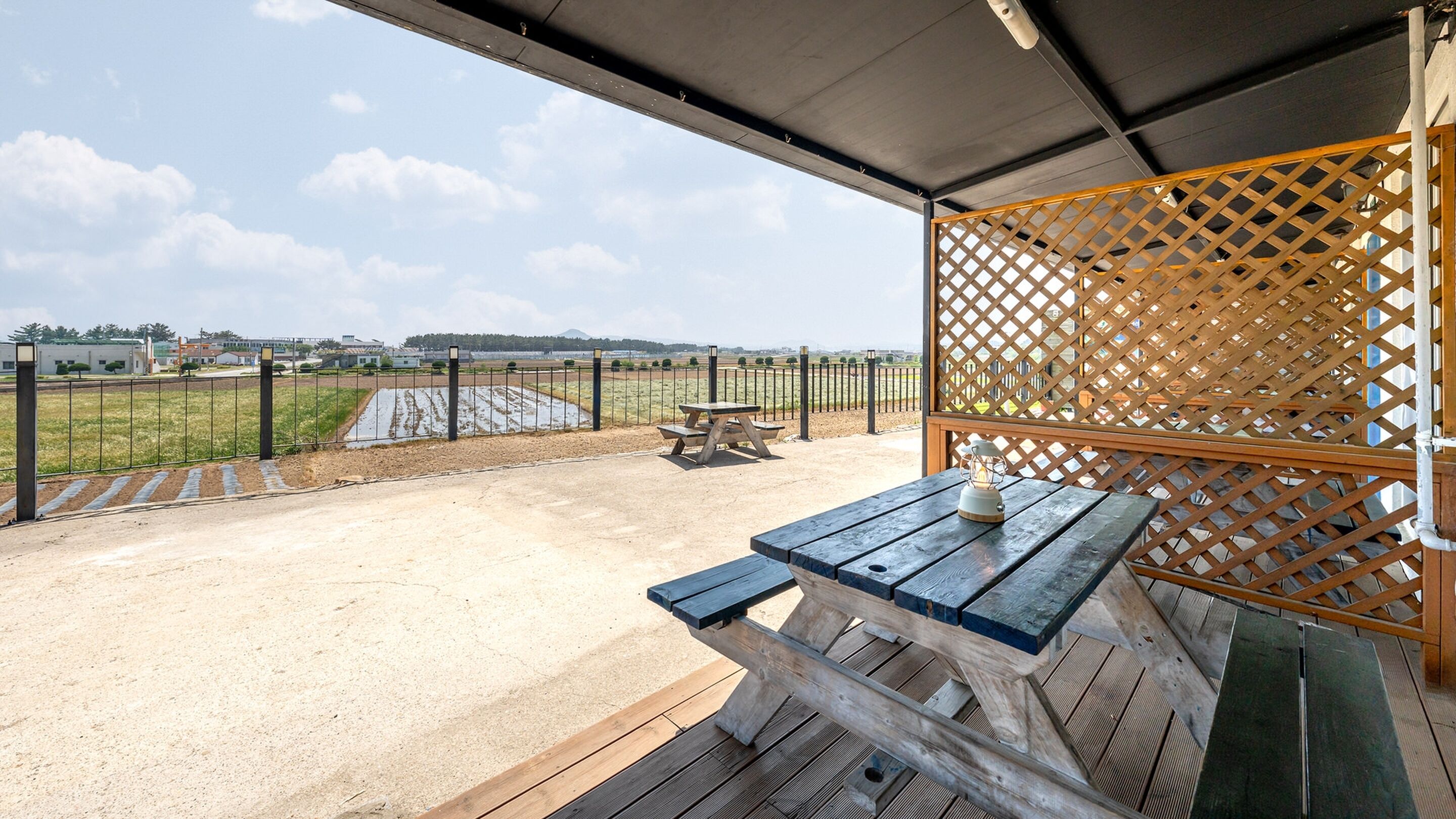 An outdoor patio area with a wooden table and benches, a picnic table, and a view of a field and distant buildings.