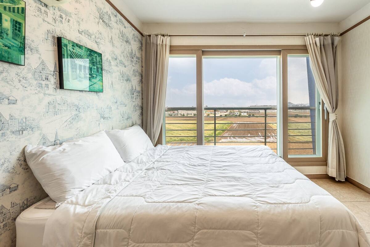 A bedroom with a large bed, a view of fields through a sliding door, and a cityscape wallpaper.