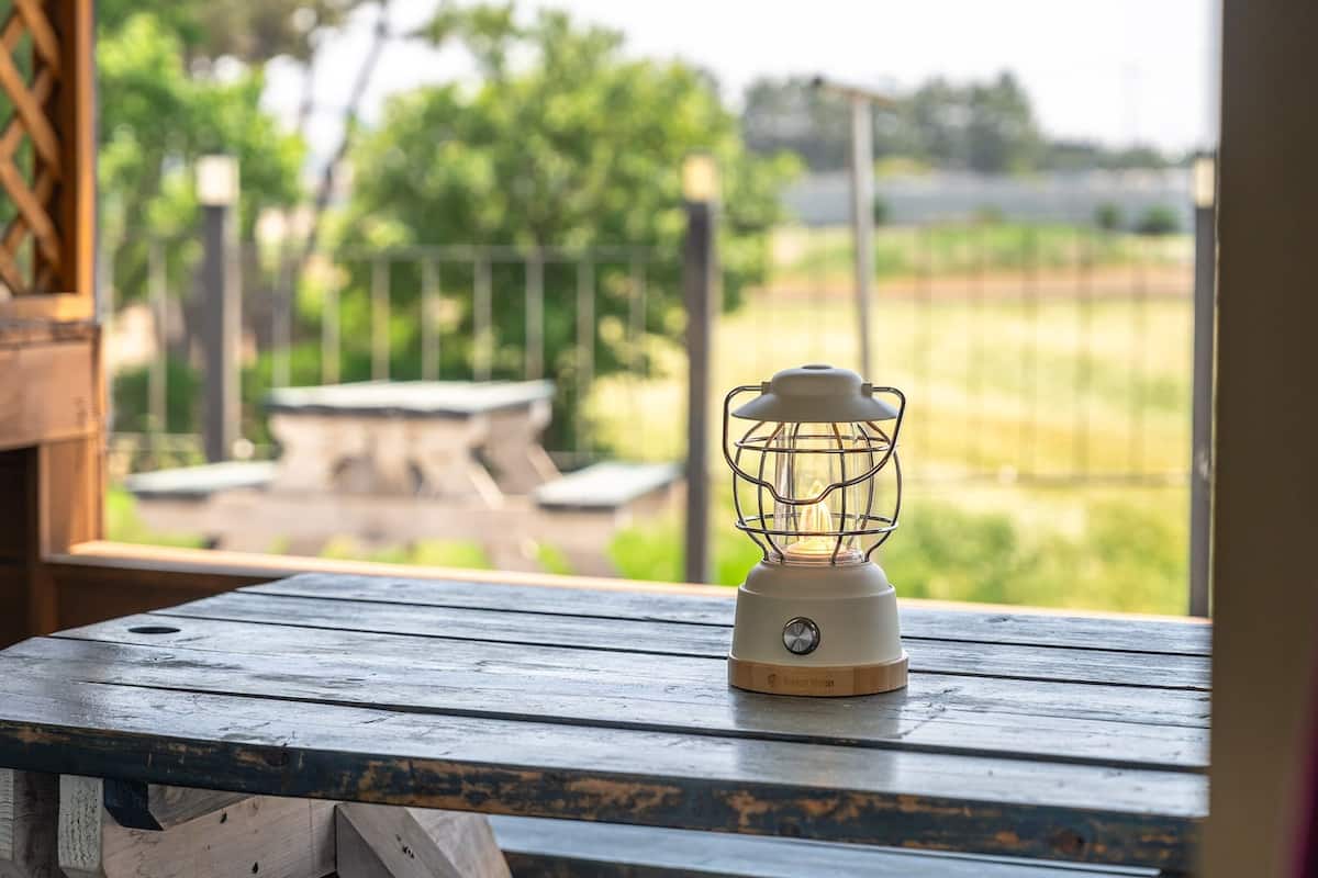 A wooden table with a lantern on it, overlooking a grassy area and a fence.