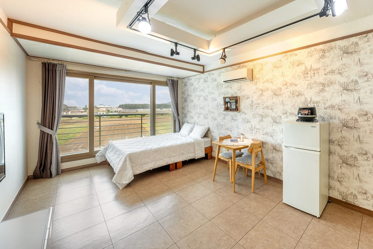 A bedroom with a bed, a desk, a chair, a refrigerator, and a window with curtains.