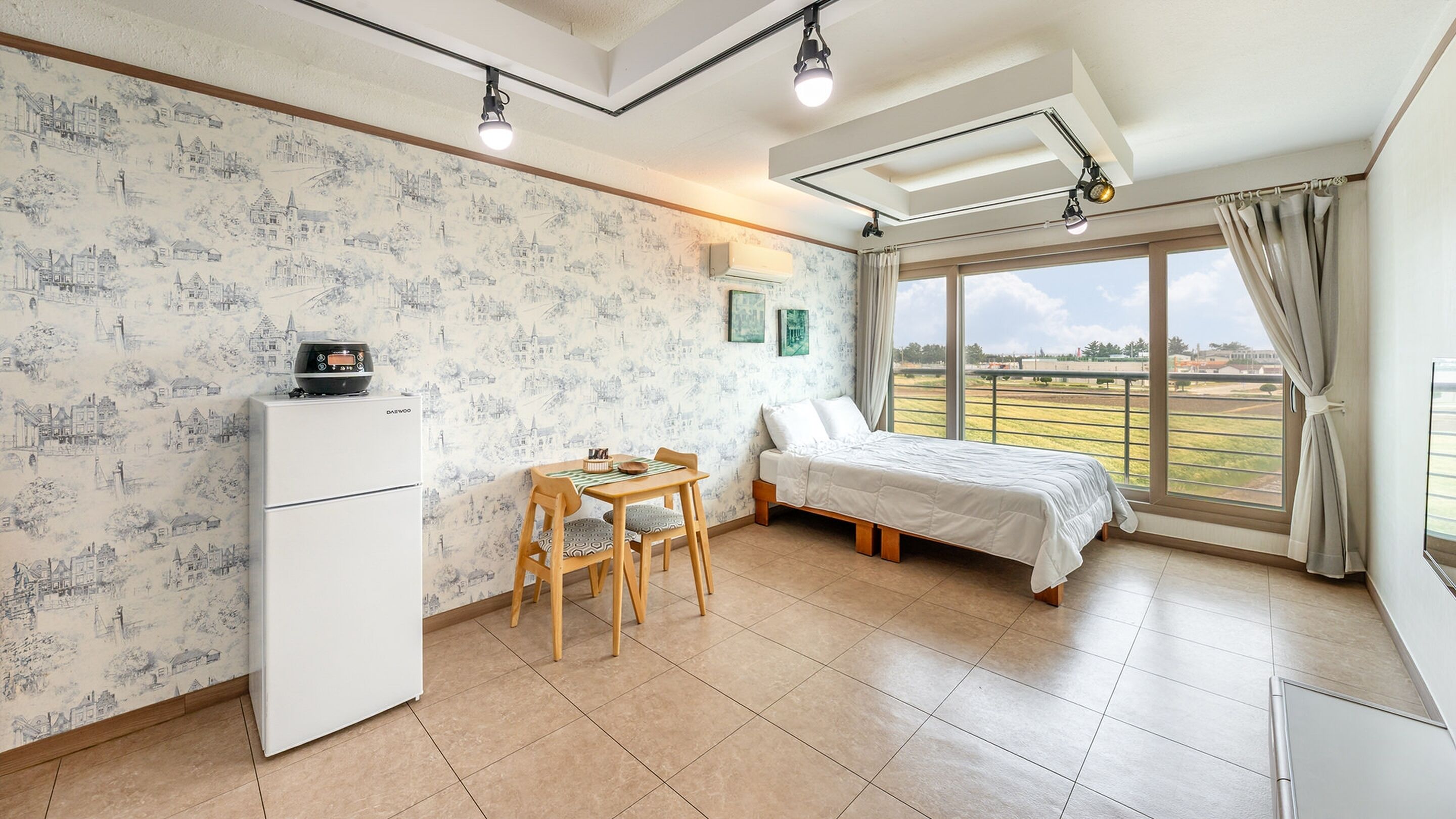 Een slaapkamer met een bed, een klein tafeltje met stoelen, een koelkast en een groot raam met uitzicht.