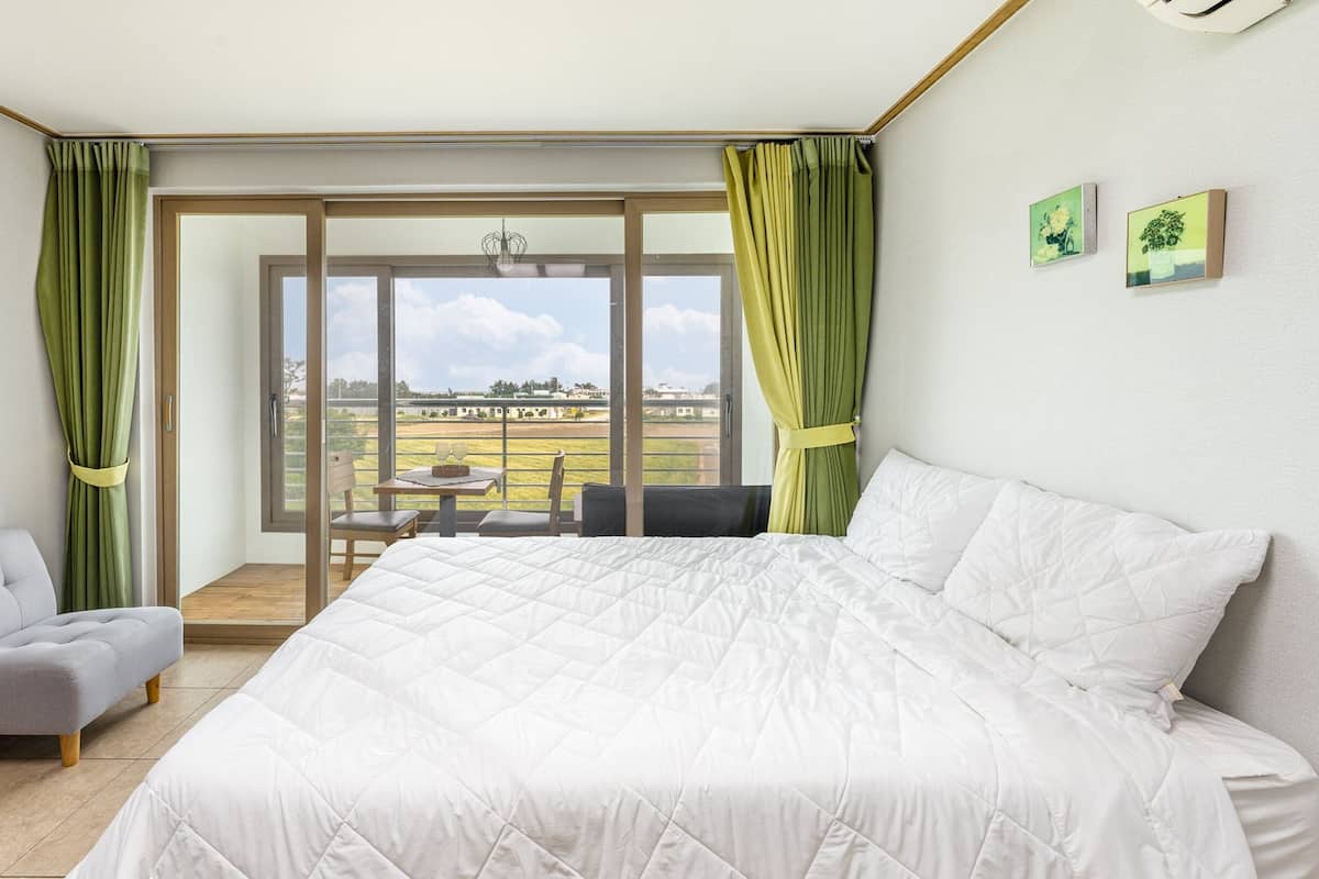 A bedroom with a large bed, a grey sofa, and a balcony with a view of the countryside.