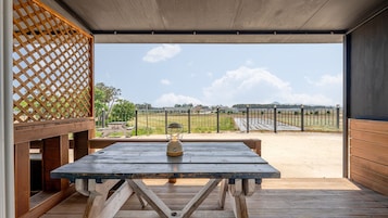 Una terraza de madera al aire libre con una mesa de picnic y un farol, con vistas a un campo vallado y edificios distantes.