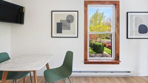 A modern room with a dining table, two chairs, a large window with a view of a garden, and two framed abstract artworks on the walls. - NY Suite | Downtown walk to Commons | Free Parking (Ithaca)