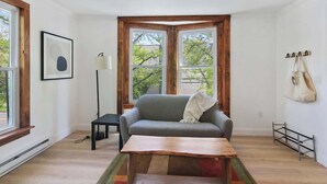 A room with a gray sofa, a wooden coffee table, a floor lamp, and a window with a view of trees. - NY Suite | Downtown walk to Commons | Free Parking (Ithaca)