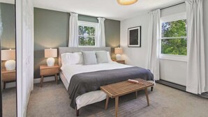 Un dormitorio con una cama grande, dos mesitas de noche con lámparas, un banco y dos ventanas con cortinas.