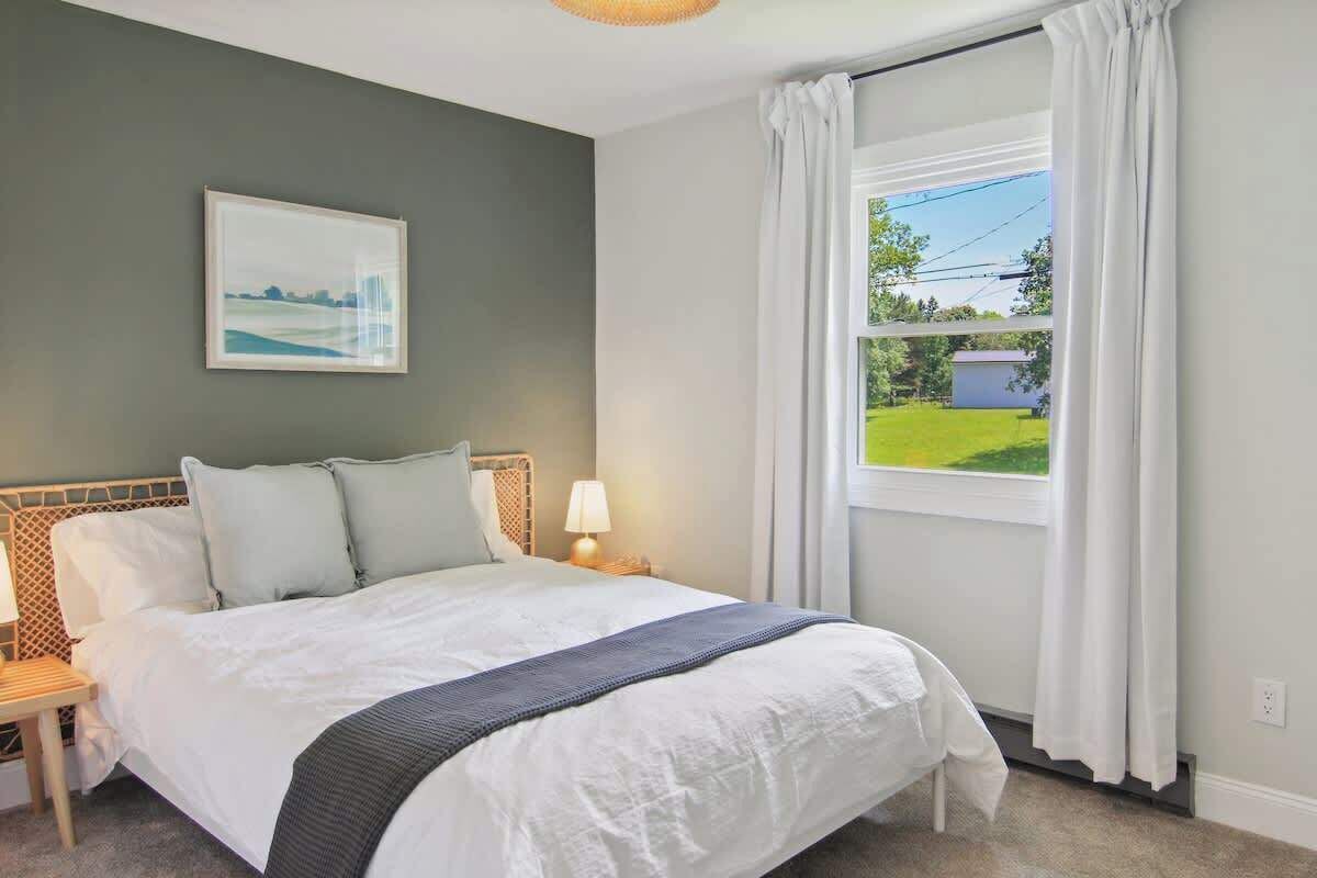 A bedroom with a large bed, a nightstand, a lamp, a window with curtains, and a painting on the wall.