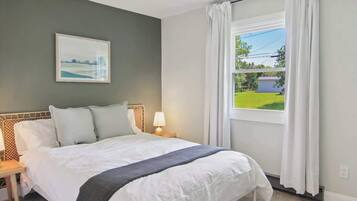 A bedroom with a large bed, a nightstand, a lamp, a window with curtains, and a painting on the wall.