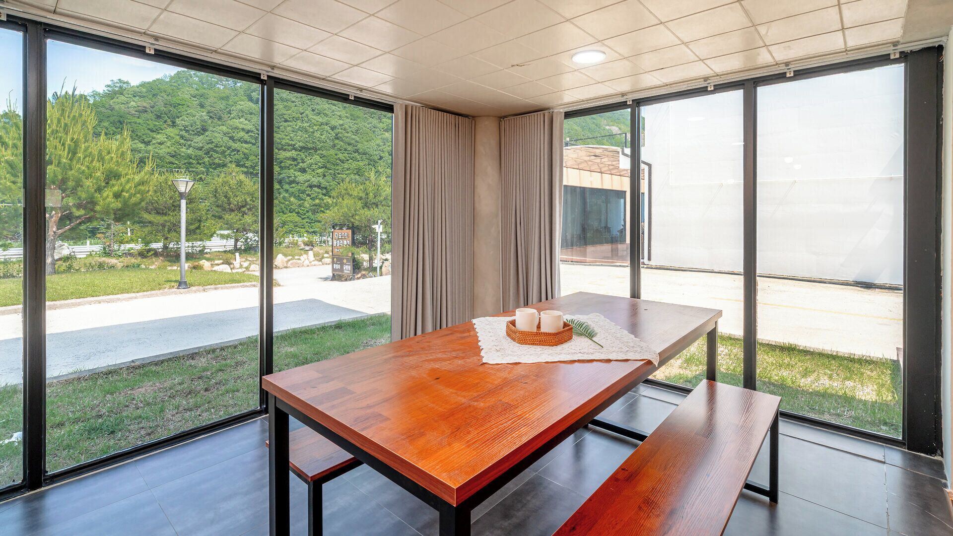 A modern dining area with a wooden table, benches, and a view of a park and building through large windows.