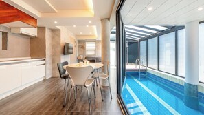 A modern hotel room with a dining area, a kitchen, and a large window overlooking a swimming pool.