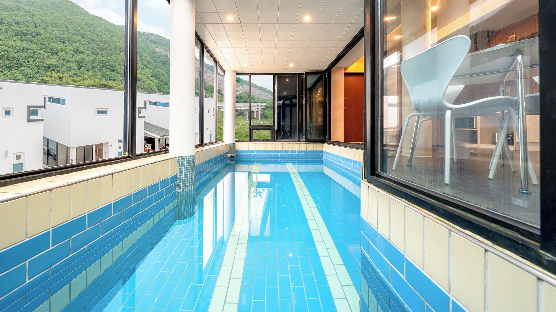Una piscina interior con suelo y paredes de azulejos, amplias ventanas y vistas a una ladera verde.