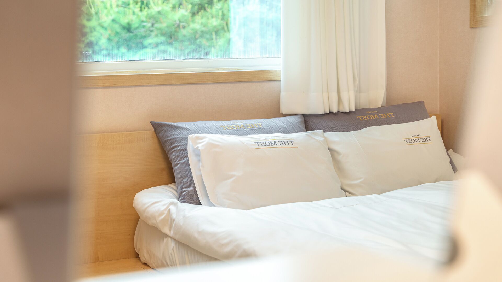 A bed with white bedding and two pillows with 'THE MYST' text, a window with a view of greenery, and a curtain.