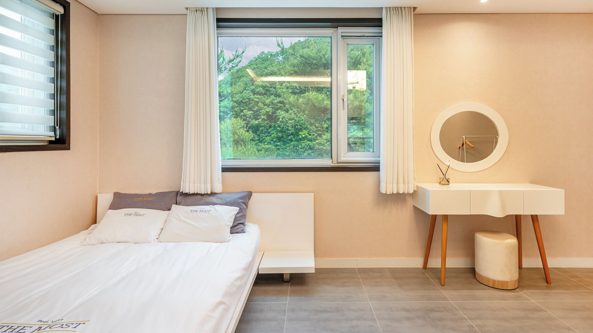 Une chambre à coucher avec un lit, une table de chevet dotée d’un miroir, une fenêtre avec des rideaux et une vue sur la verdure à l’extérieur.