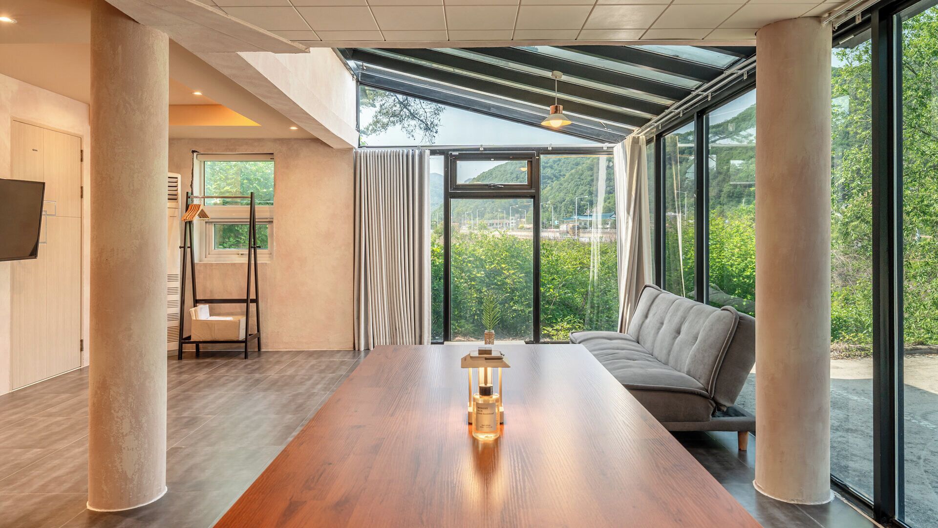 A modern interior with a long wooden table, a sofa, and large windows offering a view of greenery.
