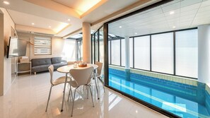 Une chambre d’hôtel moderne dotée d’une grande fenêtre donnant sur une piscine, d’un canapé, d’une table à manger avec des chaises et d’un téléviseur à écran plat fixé au mur.