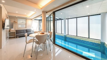 Habitación de hotel moderna con un ventanal grande con vistas a una piscina, un sofá, una mesa de comedor con sillas y un televisor de pantalla plana montado en la pared.