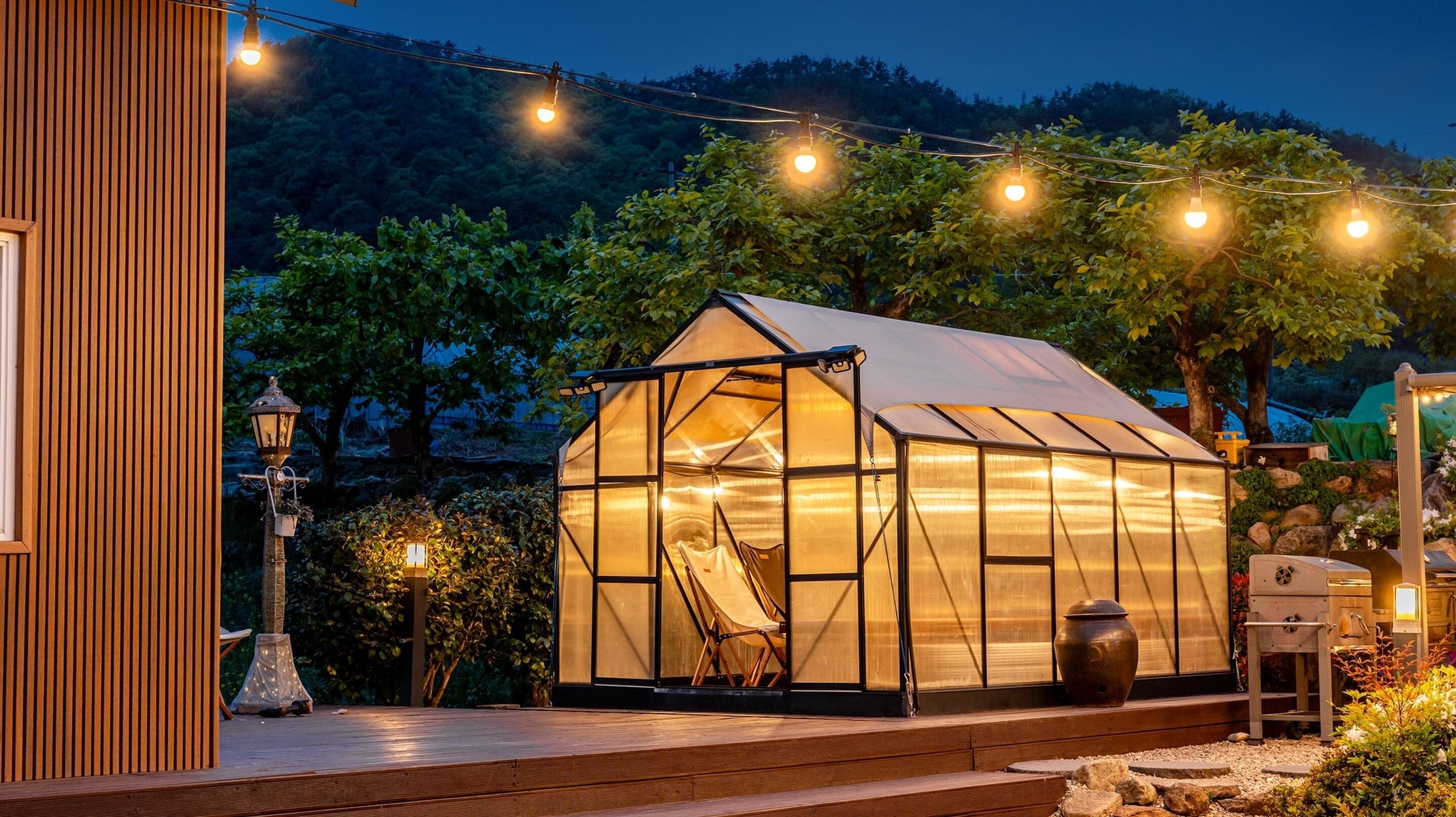 A greenhouse structure with a transparent roof, a chair inside, and a wooden deck with string lights.