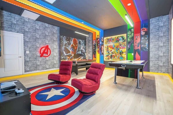 Una habitación con mesa de billar, dos sillas rojas, un tapete con temática de Capitán América y carteles de cómics en las paredes.