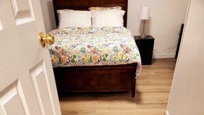 Une chambre avec un plancher en bois, un lit recouvert d’une couvre-lit à motifs floraux, une table de chevet avec une lampe et une porte dotée d’une poignée en laiton.