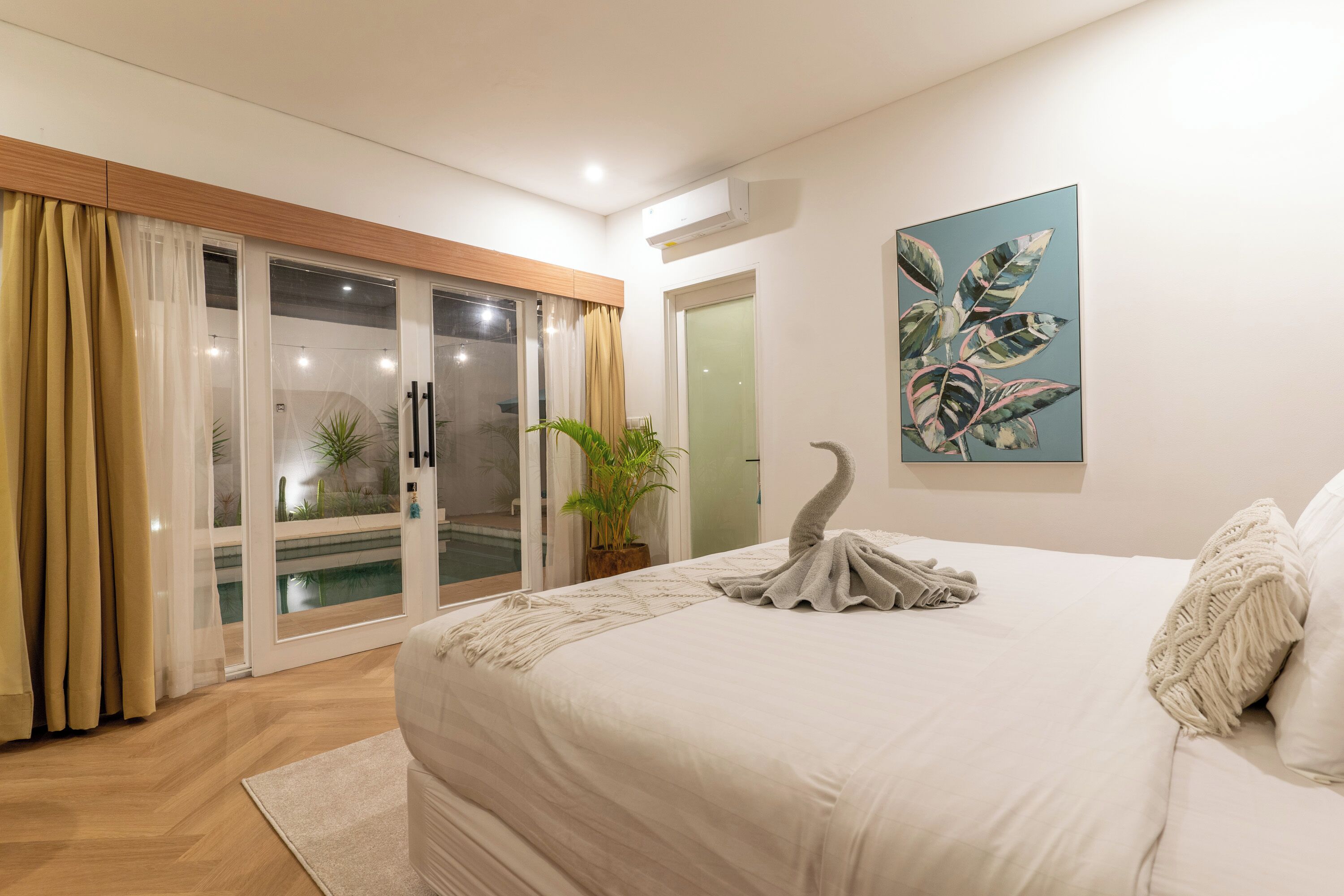 A bedroom with a large bed, a sliding glass door leading to a balcony, a potted plant, and a wall-mounted air conditioner.