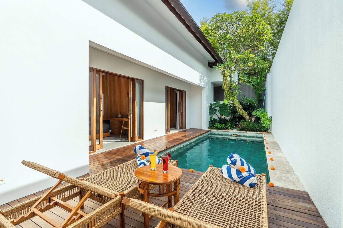 Zona de piscina con un deck de madera, sillas de descanso, una mesa pequeña con bebidas y vistas a un jardín.