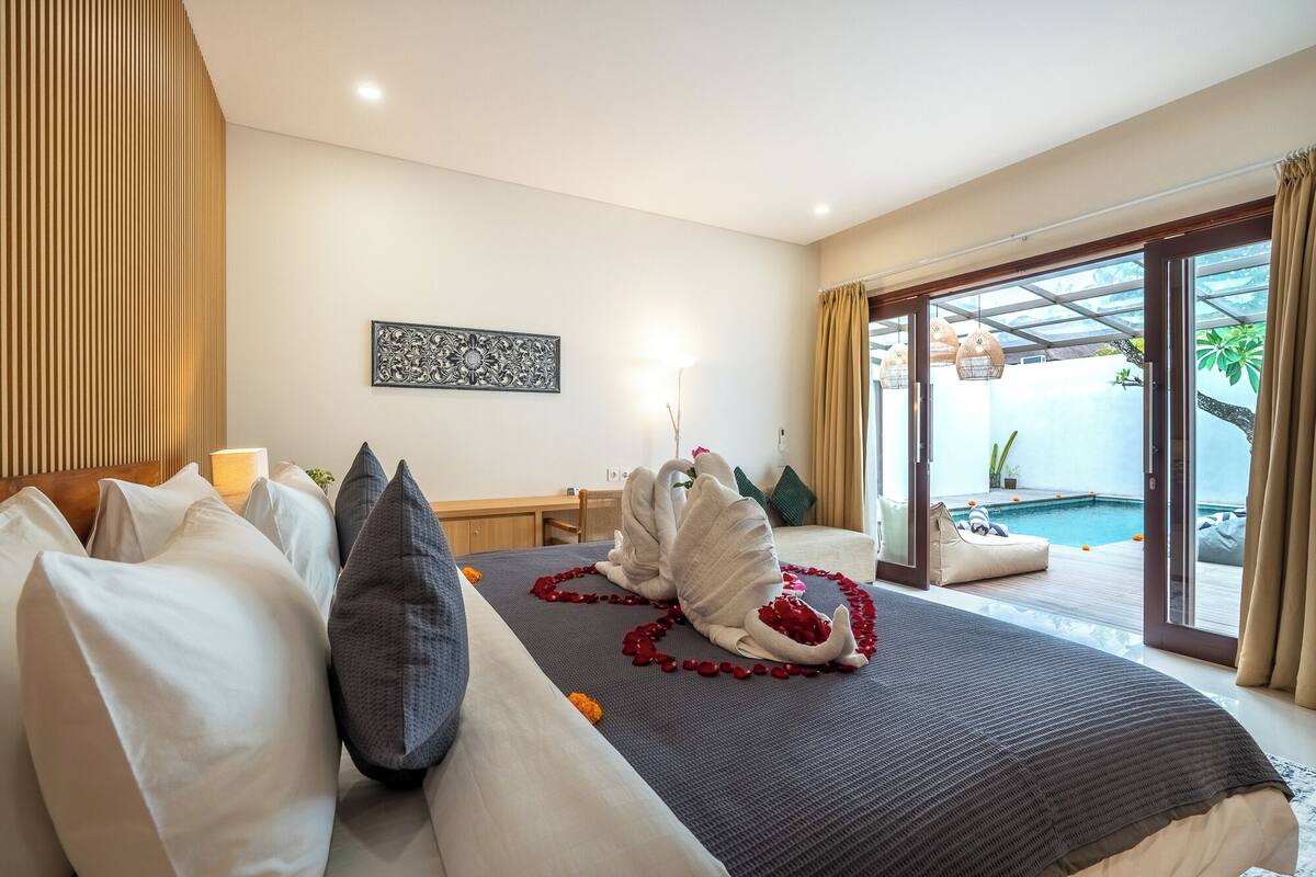 Un dormitorio con cama, almohadas, una mesita de noche, un adorno en la pared y vistas a una piscina exterior.