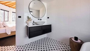 A modern bathroom with a black and white checkered floor, a round mirror above a white sink, and a wooden side table.