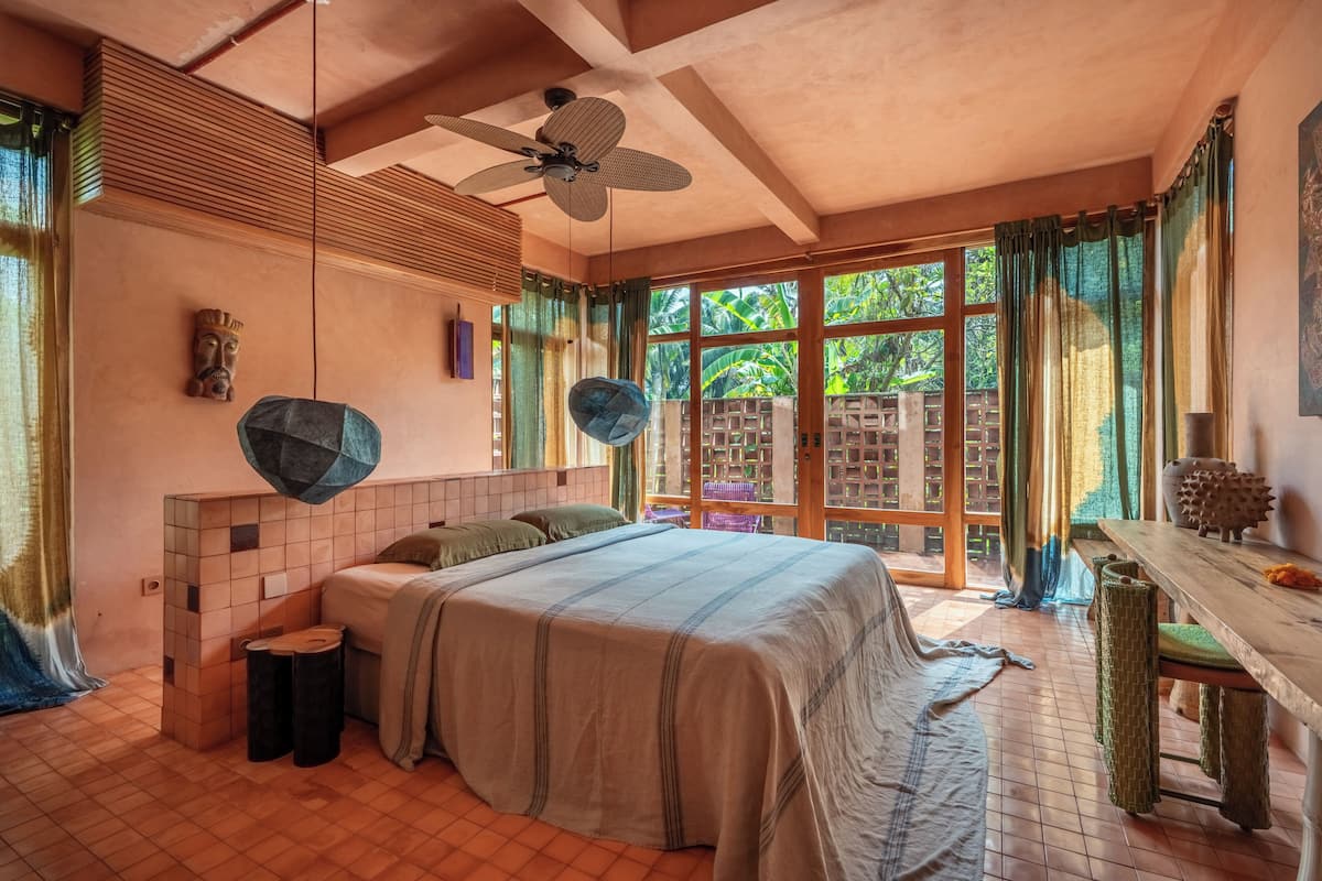 A bedroom with a bed, a ceiling fan, a window with curtains, and a view of greenery outside.