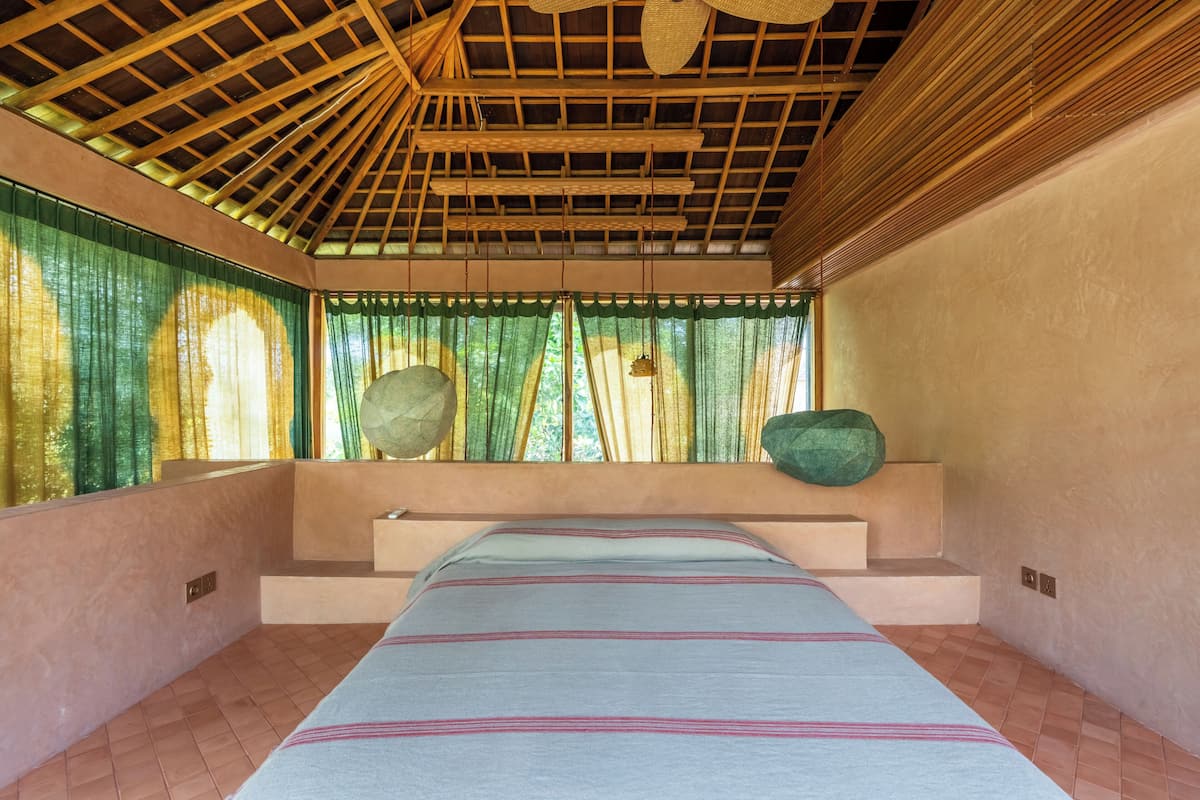 A room with a bed, a window with green curtains, a wooden ceiling, and a stone wall.