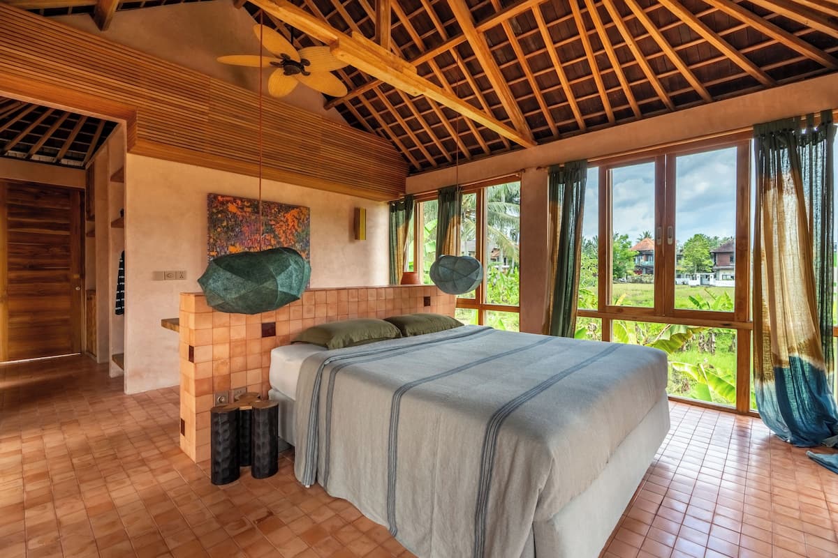 A bedroom with a large bed, wooden ceiling beams, a ceiling fan, and a view of greenery outside.