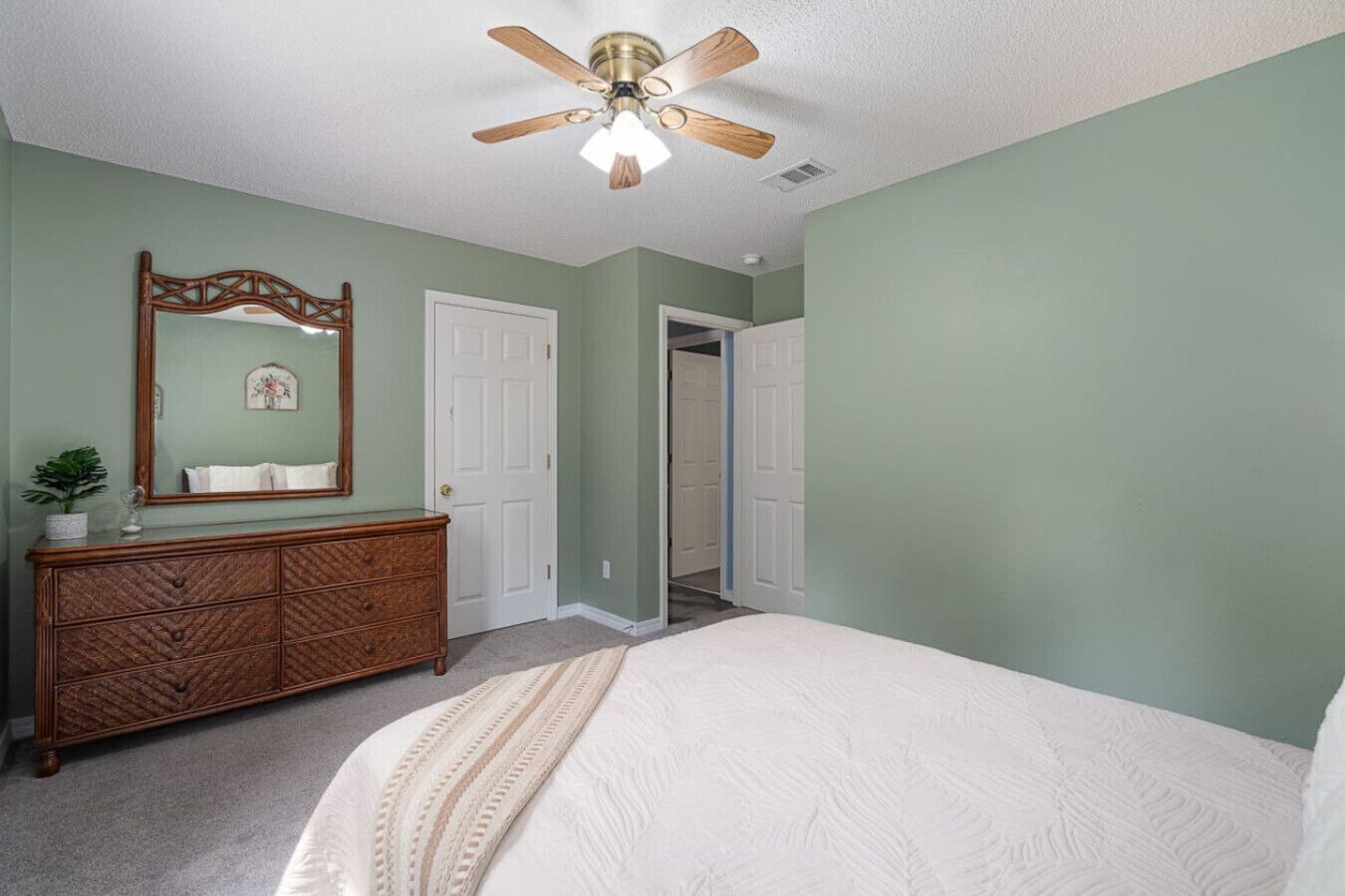 A bedroom with a bed, dresser, mirror, ceiling fan, and a door leading to another room.
