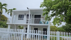 A two-story white building with balconies, a white fence, and trees in the background. - Kick back and relax in this calm, stylish space. (Ponce)