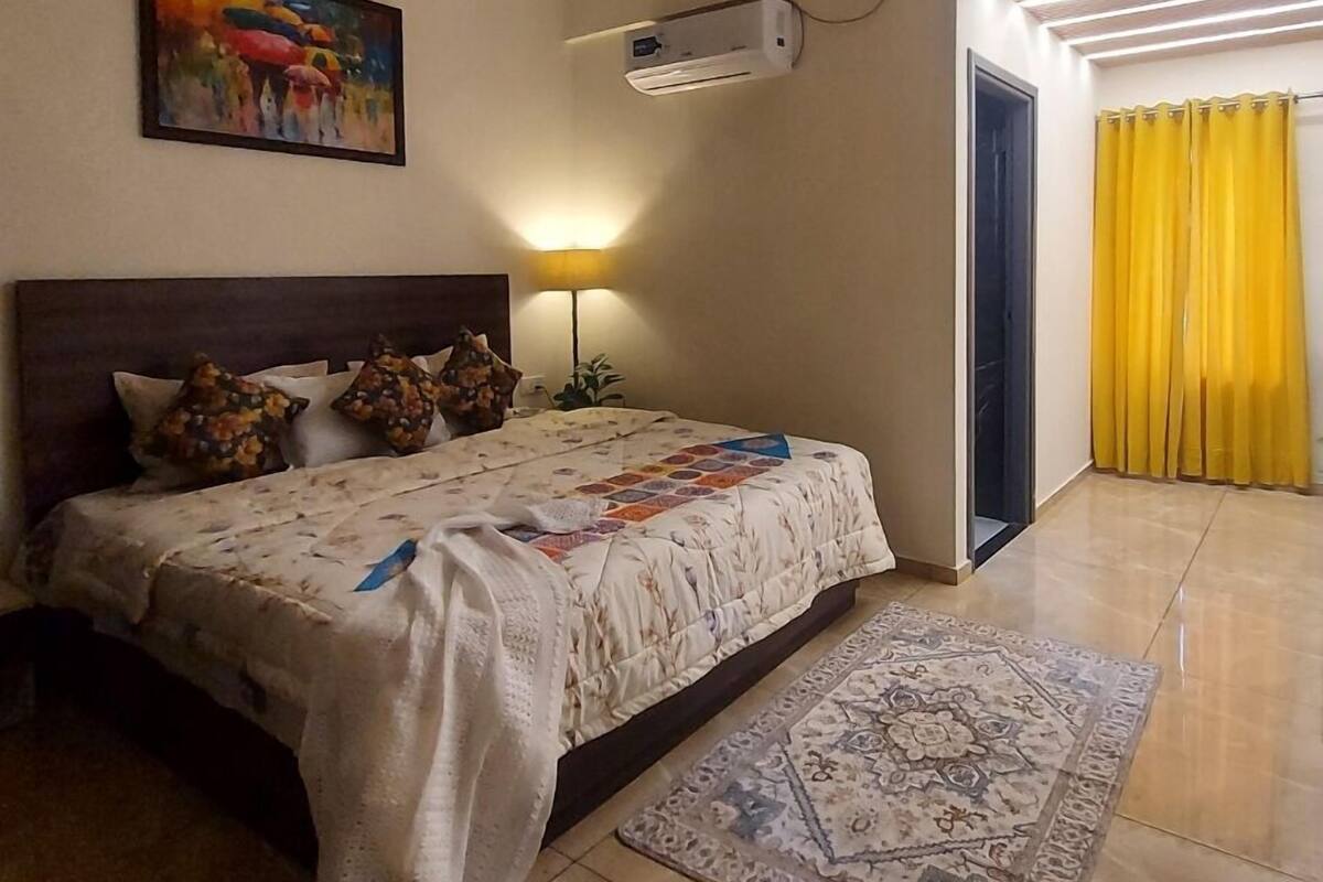 A bedroom with a bed, a nightstand, a lamp, an air conditioning unit, a painting, and a yellow curtain.