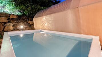 A rectangular outdoor swimming pool with a clear blue water surface, surrounded by a stone wall and a covered area.