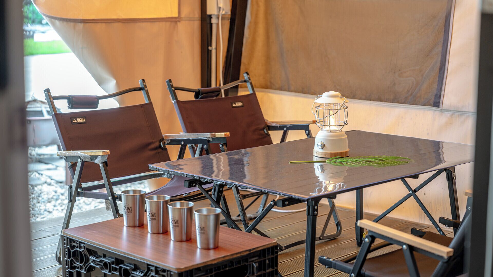 A table with a metal frame, four chairs with brown cushions, and a white table lamp.