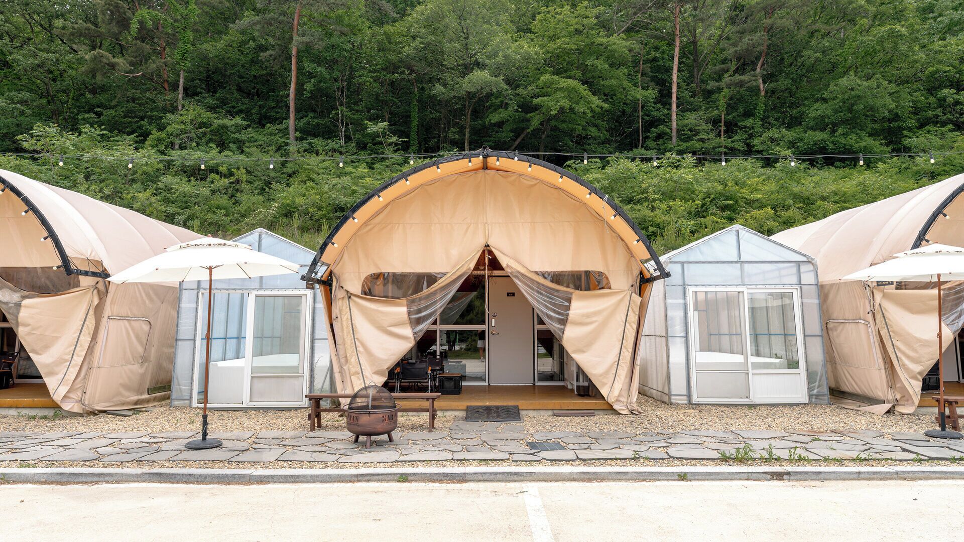 A row of tents with a central entrance, a fire pit, and a greenhouse structure.