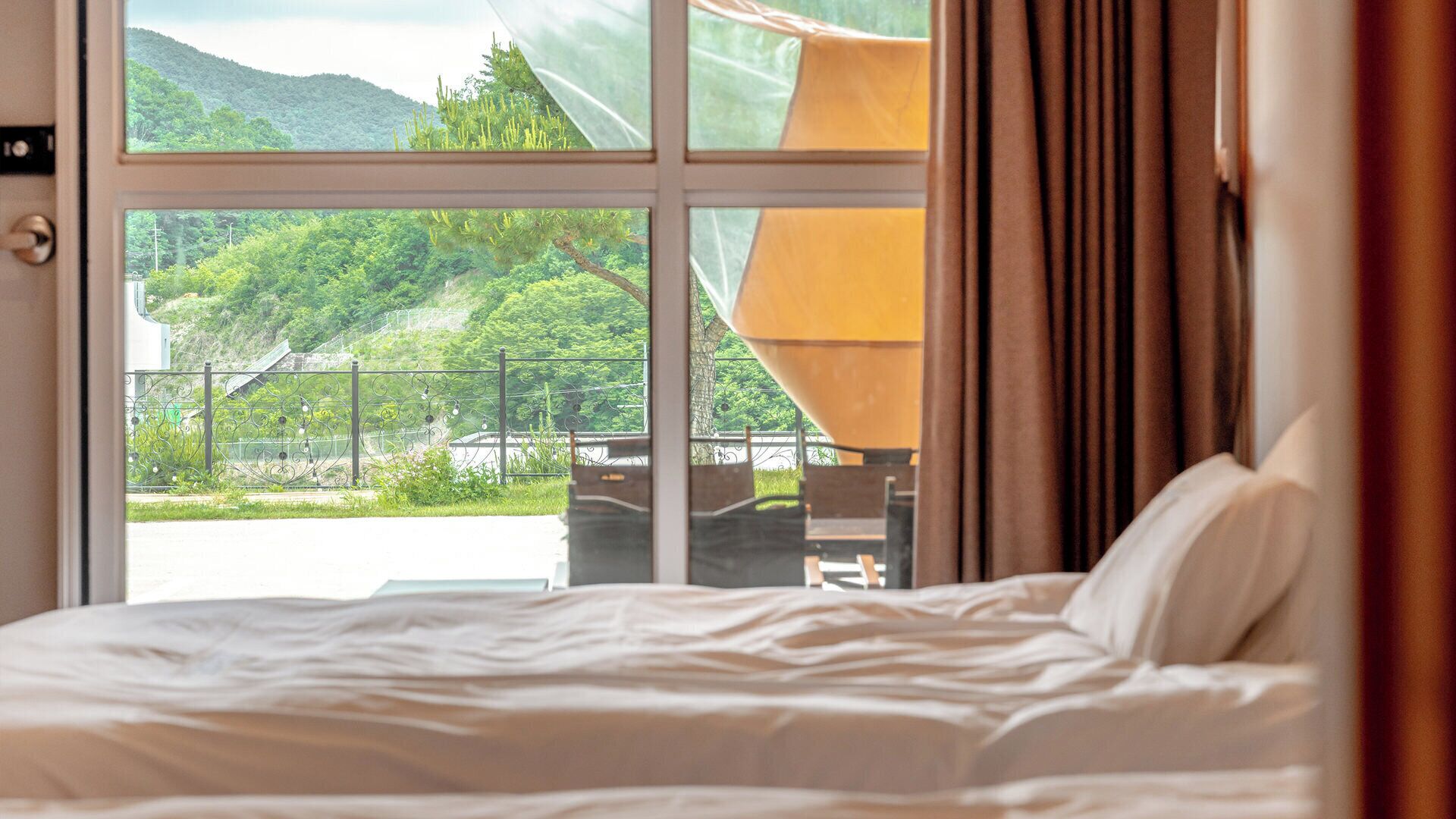 Una habitación de hotel con una cama, un ventanal con vistas a un paisaje verde y una lámpara.