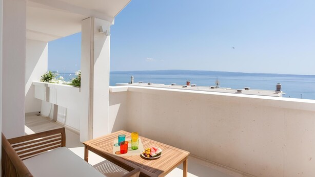A balcony with a view of the sea, a table with drinks and fruit, and a chair.