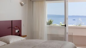 A hotel room with a bed, a nightstand, a lamp, and a view of the sea through a large window.