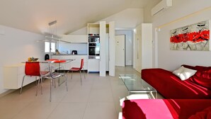 A modern living room with a red sofa and glass coffee table, a dining area with a red chair, and a kitchen with white cabinets and a microwave.