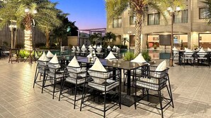 An outdoor dining area with tables and chairs, palm trees, and a building in the background. - The Imperial Prime (Bhilwara)