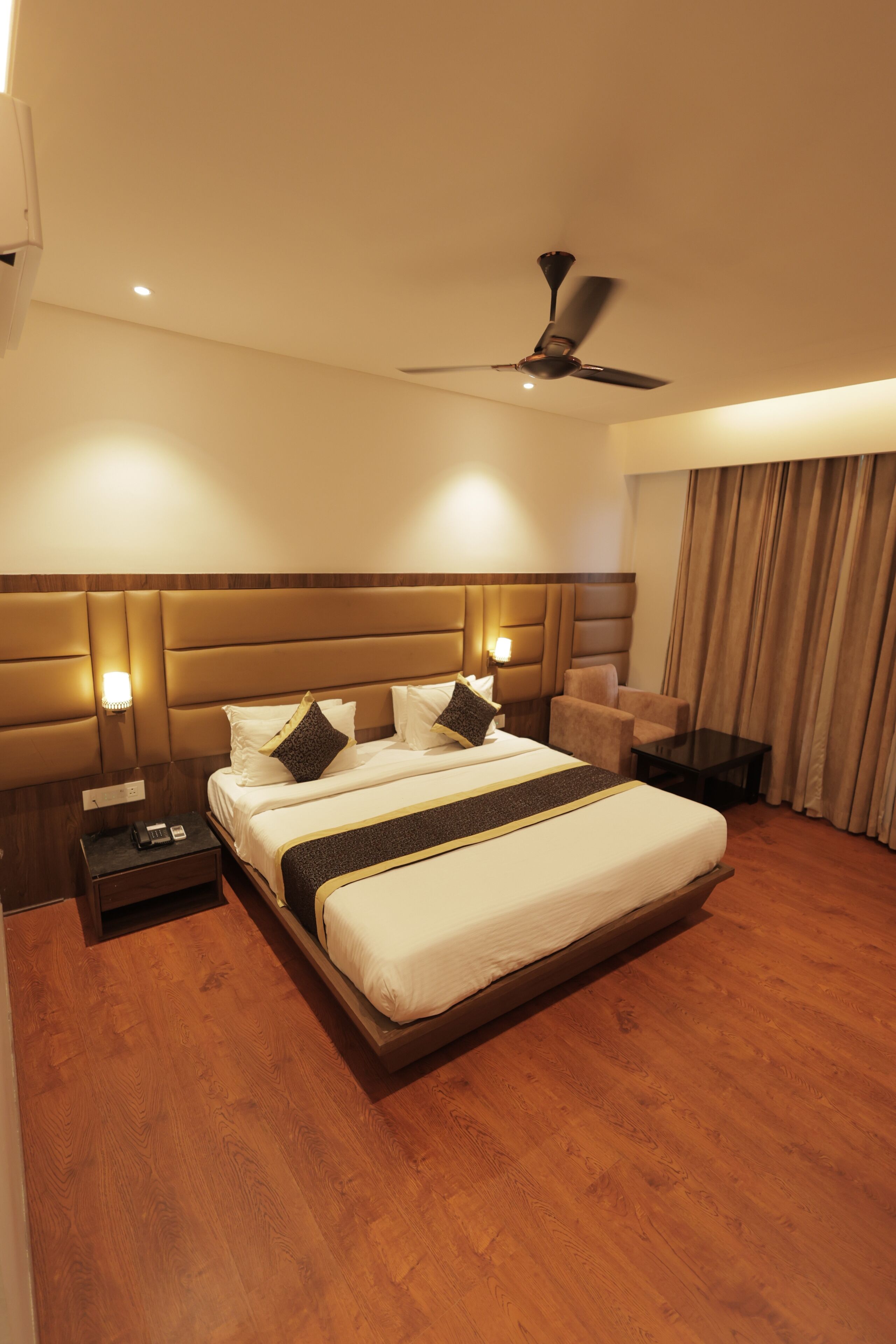 A hotel room with a large bed, a nightstand, a desk, a chair, a ceiling fan, and a window with curtains.