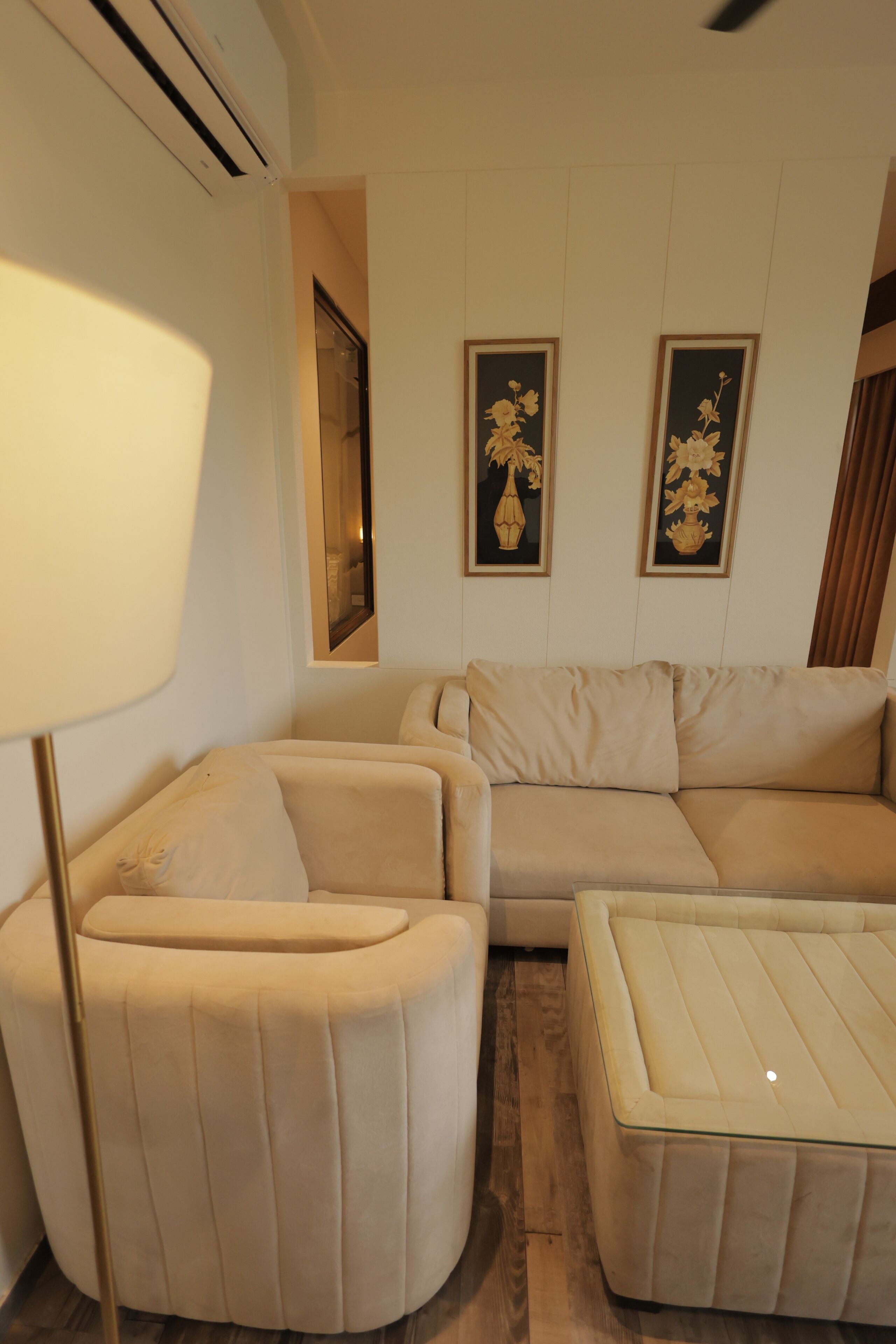 Un salon comprenant un canapé, un fauteuil et un pouf, une lampe sur pied, ainsi que deux tableaux floraux encadrés accrochés au mur.